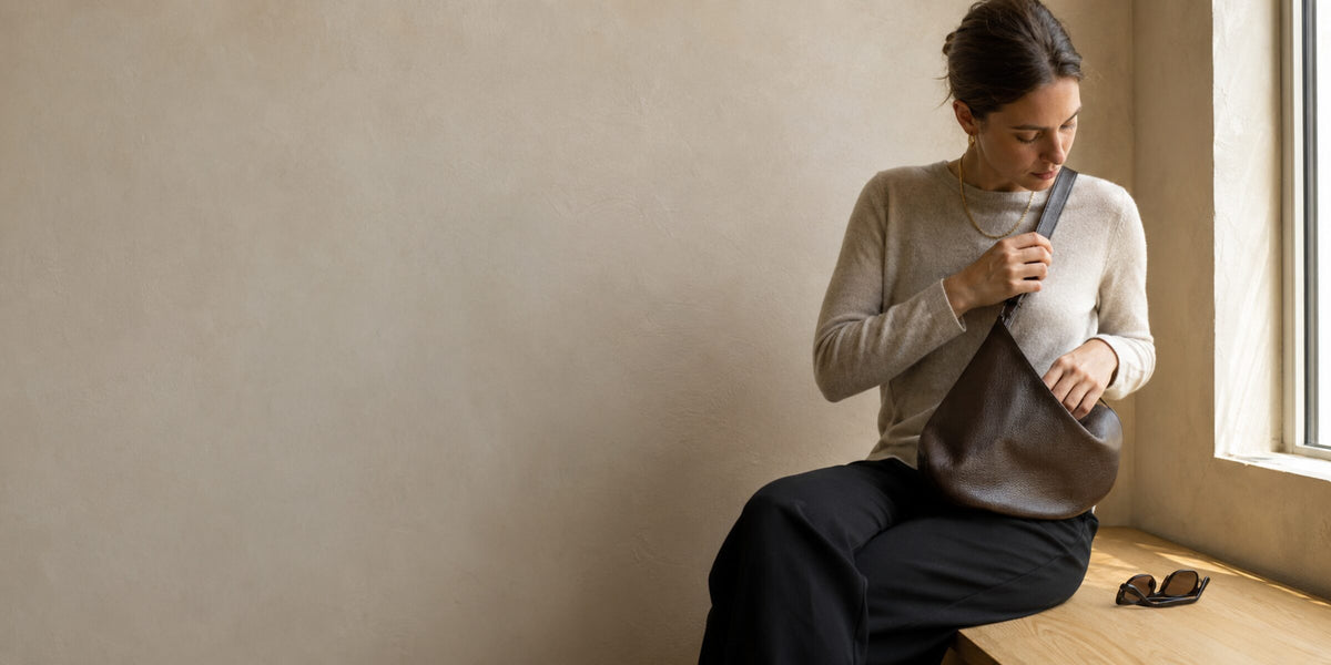 GLOSHWEAR Fall 2026 — woman in a bone knit wearing a chocolate leather crescent bag on a sunlit plaster bench