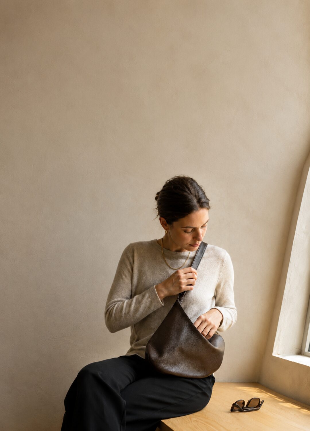 GLOSHWEAR Fall 2026 — woman in a bone knit wearing a chocolate leather crescent bag on a sunlit plaster bench
