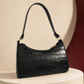 A black crocodile-embossed leather shoulder bag with gold-tone hardware, displayed on a cream-colored pedestal against a beige and dark red background.