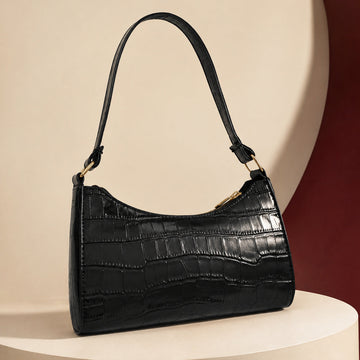 A black crocodile-embossed leather shoulder bag with gold-tone hardware, displayed on a cream-colored pedestal against a beige and dark red background.