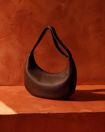 Dark chocolate crescent shoulder bag on a deep terracotta plaster ledge