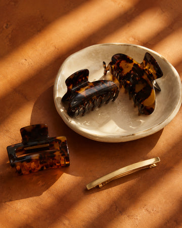 Tortoiseshell claw clips and a gold barrette in dappled light on ochre clay