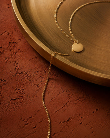 Fine gold pendant necklace draped over a brushed brass tray on terracotta