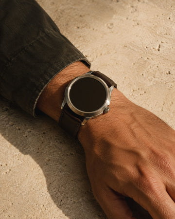 Plain steel watch with brown leather strap on a wrist over pale stone